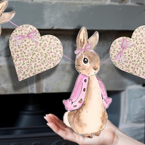 Può includere: Un coniglio rosa e marrone con un fiocco rosa e una giacca rosa fa parte di una ghirlanda con cuori floreali rosa e verdi.