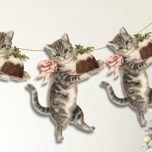 Può includere: Ghirlanda di tre sagome di gatti tabby grigi, ognuno con un budino marrone su un piatto. I gatti indossano fiocchi rosa e decorazioni di agrifoglio verde. La ghirlanda è appesa a un cordoncino sottile di colore naturale.