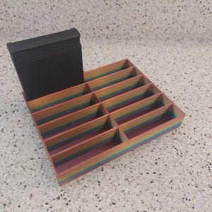 May include: A rainbow-colored organizer with multiple compartments for storing small items. A black rectangular box sits on top of the organizer.