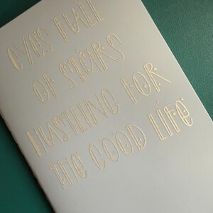 May include: A light gray notebook with gold lettering. The text reads "Eyes full of stars hustling for the good life." The notebook is on a green surface.