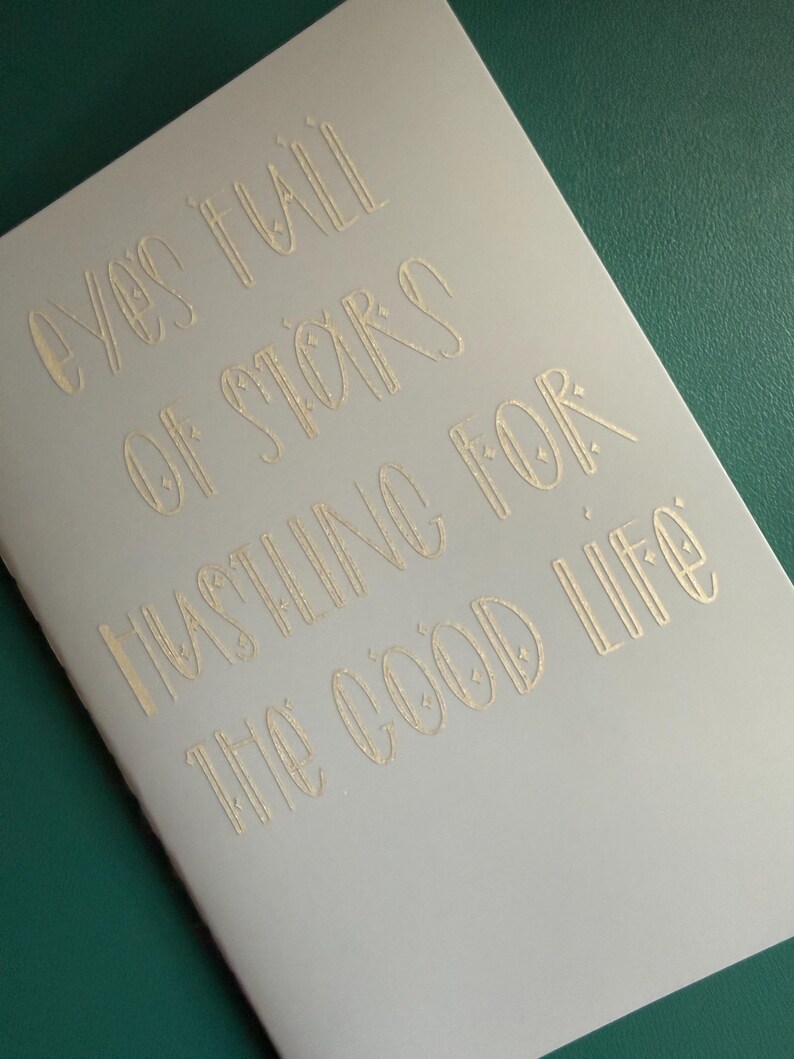 May include: A light gray notebook with gold lettering. The text reads "Eyes full of stars hustling for the good life." The notebook is on a green surface.