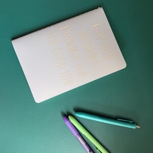 May include: A white notebook with gold lettering that reads "OVER HALF OF STARS HASTING FOR THE GOOD LIFE" is positioned on a teal surface. Three pens in purple, green, and teal are placed below the notebook, offering a colorful contrast.
