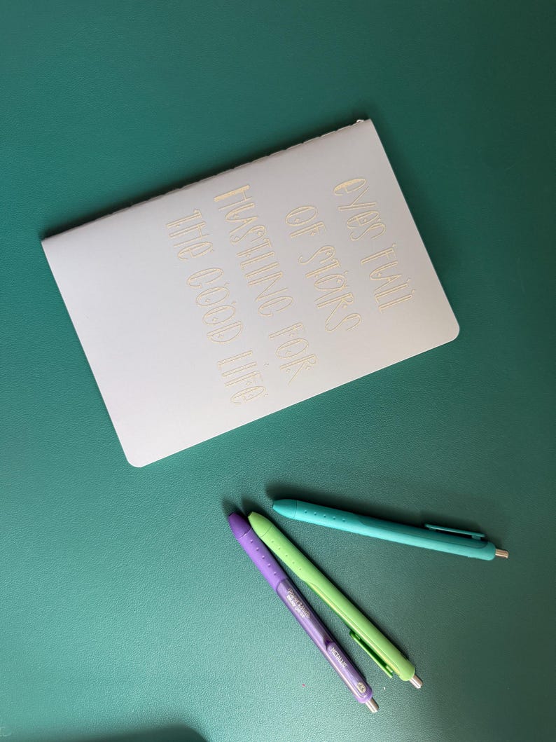 May include: A white notebook with gold lettering that reads "OVER HALF OF STARS HASTING FOR THE GOOD LIFE" is positioned on a teal surface. Three pens in purple, green, and teal are placed below the notebook, offering a colorful contrast.