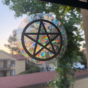 May include: A circular window decal featuring a black pentagram symbol centered on a background with a prismatic, shattered glass effect that reflects rainbow colors when light passes through the transparent adhesive material.