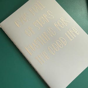 May include: A light gray notebook with gold lettering that reads "Eyes Full of Stars Hustling for the Good Life." The notebook is on a green surface. The notebook is closed and has a simple design.