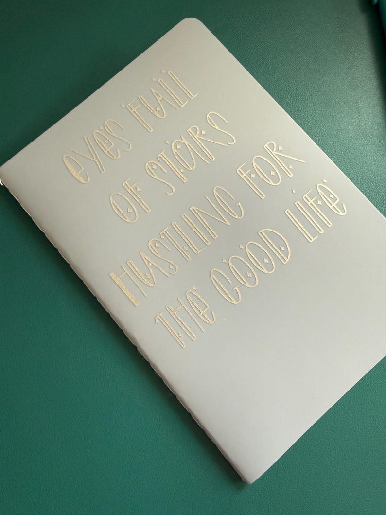 May include: A light gray notebook with gold lettering that reads "Eyes Full of Stars Hustling for the Good Life." The notebook is on a green surface. The notebook is closed and has a simple design.