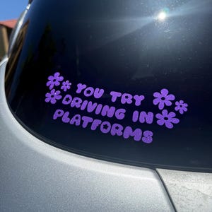May include: Purple glitter vinyl decal on a car window. The decal features the text "YOU TRY DRIVING IN PLATFORMS" in a playful font, with flower accents. The car's silver paint is visible in the foreground.