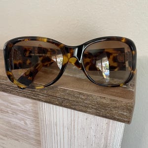 May include: Tortoiseshell sunglasses with brown lenses. The frames feature a dark brown and yellow pattern. The sunglasses have a rectangular shape and are resting on a wooden surface. The text "Oliver Goldsmith & England" is visible on the lens.
