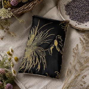 Peut inclure: Un coussin rectangulaire noir avec un motif floral doré. Il est posé sur un tissu de couleur neutre, entouré de fleurs séchées, d'un panier en osier et d'une petite assiette de lavande.