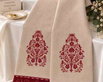 May include: A beige linen runner featuring two symmetrical red cross-stitch floral patterns and a decorative red lace trim along the bottom edges. The fabric has a visible woven texture, and the embroidery displays intricate botanical motifs.