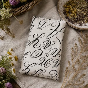 Puede incluir: Un cojín rectangular blanco roto con letras cursivas negras. El cojín está colocado sobre una superficie de lino de color neutro, rodeado de flores secas y una cesta de mimbre. La estética general es rústica y elegante.