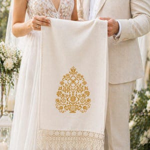 May include: A rectangular off-white fabric runner featuring a gold-coloured cross-stitch floral pattern in the centre. The bottom edge is finished with a wide, intricate cream-coloured lace trim with a scalloped design.