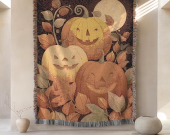May include: A woven tapestry featuring a Halloween-themed design. The artwork depicts three smiling jack-o'-lanterns in shades of orange and yellow, surrounded by autumn leaves and a full moon. The tapestry has a fringed border.