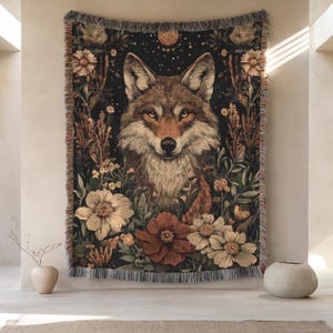 May include: A woven tapestry featuring a detailed portrait of a fox surrounded by flowers and foliage. The color palette includes shades of brown, beige, and green against a dark background. The tapestry has a fringed edge.