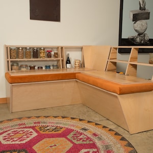May include: A custom-built, light-colored wooden corner bench with orange cushions. The bench features built-in shelves holding jars of food and a wine bottle. A colorful rug is in the foreground, and artwork hangs on the wall.
