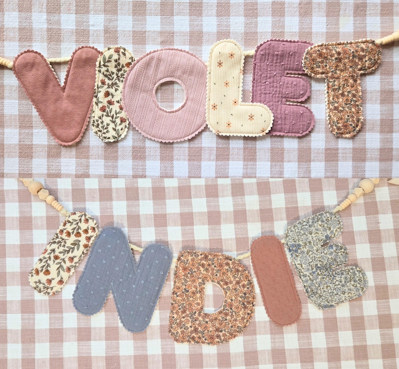 Op de afbeelding: Twee stoffen banners met de woorden "VIOLET" en "INDI". De letters zijn in verschillende kleuren en patronen, waaronder bloemenprints en effen kleuren. De banners zijn geregen op houten kralen en staan tegen een roze en witte geruite achtergrond.