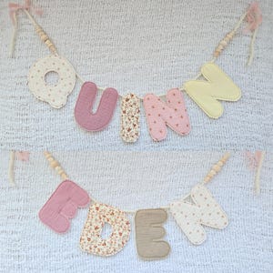 Op de afbeelding: Een handgemaakte slinger met de namen "UNN" en "EDEN". De letters zijn in roze, bloemenpatroon en cr&egrave;me, verbonden met houten kralen en roze linten. Perfect voor kinderkamer- of feestdecoratie.