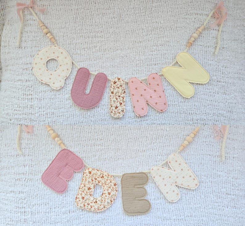 Op de afbeelding: Een handgemaakte slinger met de namen "UNN" en "EDEN". De letters zijn in roze, bloemenpatroon en cr&egrave;me, verbonden met houten kralen en roze linten. Perfect voor kinderkamer- of feestdecoratie.