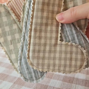 Op de afbeelding: Een verzameling handgemaakte stoffen pads in verschillende patronen en kleuren. De pads hebben geschulpte randen en zijn gemaakt van materialen met ruit- en streepmotieven. Het kleurenpalet omvat tinten beige, grijs en roze.