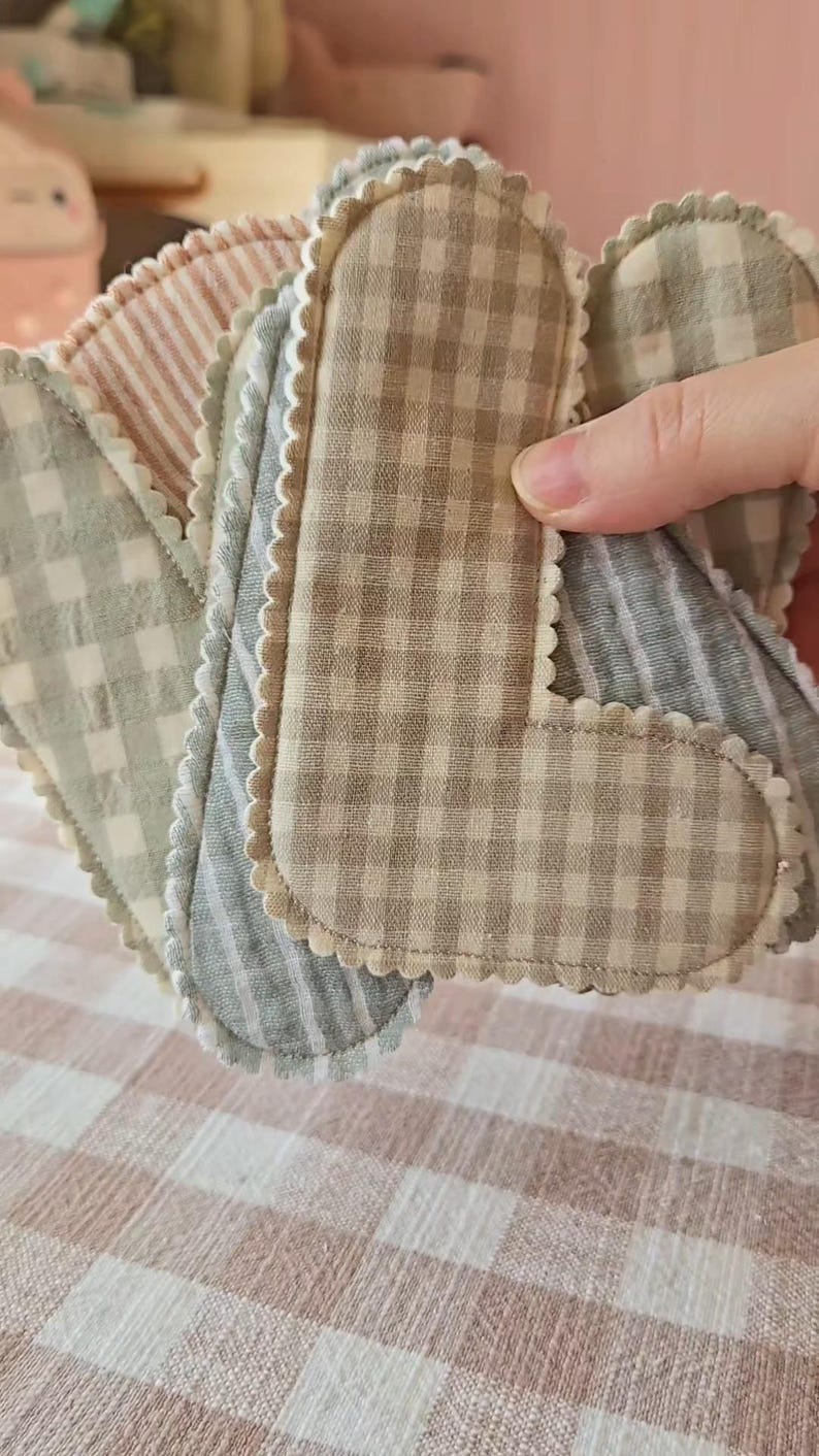 Op de afbeelding: Een verzameling handgemaakte stoffen pads in verschillende patronen en kleuren. De pads hebben geschulpte randen en zijn gemaakt van materialen met ruit- en streepmotieven. Het kleurenpalet omvat tinten beige, grijs en roze.