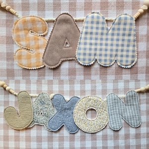 Op de afbeelding: Twee stoffen naamvlaggen met de namen "SAM" en "JASON". De letters zijn in verschillende patronen en kleuren, waaronder ruitjes, bloemen en strepen. Elke letter is omrand met een geschulpte rand en geregen op een houten kralen slinger.