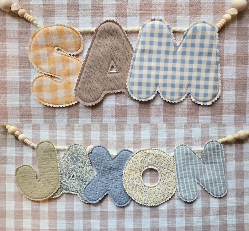 Op de afbeelding: Twee stoffen naamvlaggen met de namen "SAM" en "JASON". De letters zijn in verschillende patronen en kleuren, waaronder ruitjes, bloemen en strepen. Elke letter is omrand met een geschulpte rand en geregen op een houten kralen slinger.