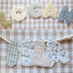 Op de afbeelding: Een handgemaakte stoffen banner met de namen "LOGAN" en "ISLA" uitgespeld in individuele, decoratieve letters. De letters zijn in verschillende kleuren en patronen, waaronder bloemen-, streep- en ruitmotieven, en zijn op een neutraal gekleurde stof genaaid.