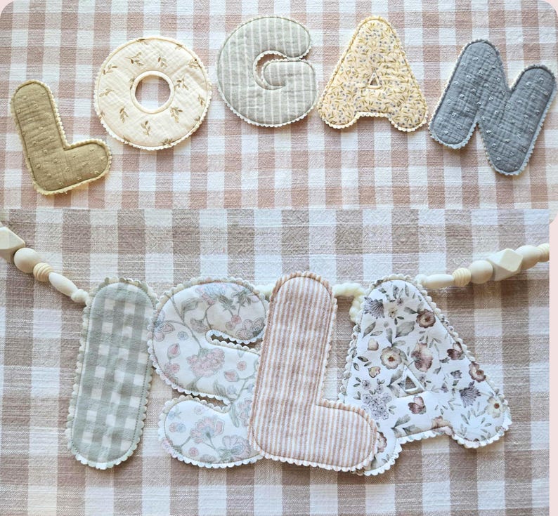 Op de afbeelding: Een handgemaakte stoffen banner met de namen "LOGAN" en "ISLA" uitgespeld in individuele, decoratieve letters. De letters zijn in verschillende kleuren en patronen, waaronder bloemen-, streep- en ruitmotieven, en zijn op een neutraal gekleurde stof genaaid.