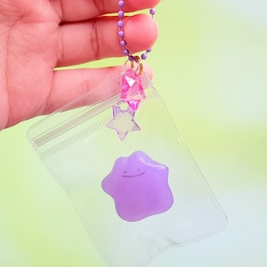 May include: A clear plastic keychain with a purple star-shaped charm inside. The keychain has a pink star charm and a pink crystal charm attached to it.