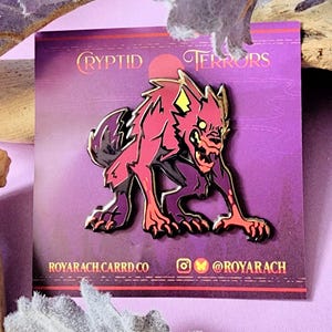 Könnte beinhalten: Eine rote und schwarze Emaille-Anstecknadel mit einem knurrenden, rotäugigen, wolfartigen Wesen mit leuchtend gelben Augen. Die Anstecknadel befindet sich auf einem violetten Hintergrund mit dem Text "CRYPTID TERRORS" in goldenen Buchstaben. Der Text "ROYARACHCARRD.CO" und "@ROYARACH" ist ebenfalls in goldenen Buchstaben sichtbar.