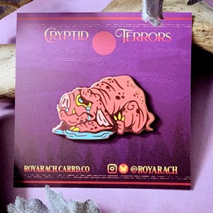 May include: A pink and yellow enamel pin featuring a cartoon monster with a sad expression. The pin is on a purple card with the text "Cryptid Terrors" and "Royarachcarrd.co" and "@royarach".