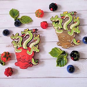 Puede incluir: Dos pines de esmalte que representan una hidra de múltiples cabezas, una con un esquema de color rojo y verde y la otra con un esquema de color dorado y verde. Ambos pines se muestran sobre un fondo de madera blanco.