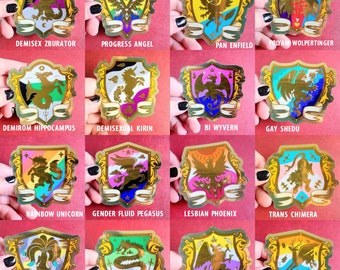 May include: A collection of sixteen shield-shaped stickers with gold foil accents. Each sticker features a unique design with a mythical creature and text, including "DEMISEX ZBURATOR," "PROGRESS ANGEL," and "RAINBOW UNICORN." The stickers are held by a person with black nail polish.