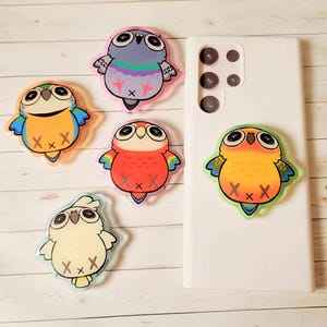 May include: Five bird-themed phone grips featuring cartoon illustrations of a pigeon, a yellow and blue parrot, a red parrot, a white cockatoo, and an orange parrot, with one grip attached to a white rectangular smartphone case.