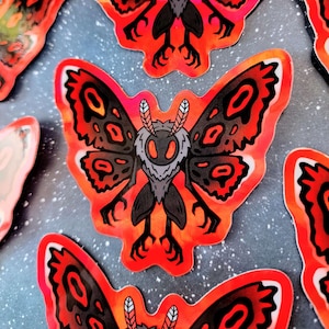May include: A set of four stickers featuring a black and red moth with large wings and a menacing expression. The moth is set against a dark blue background with white stars.
