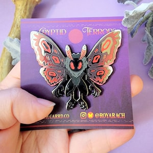 May include: A black and silver moth pin with red and tan wing details. The pin is on a purple card with the words "Cryptid Terrors" and social media handles. The moth has large eyes and a detailed body.