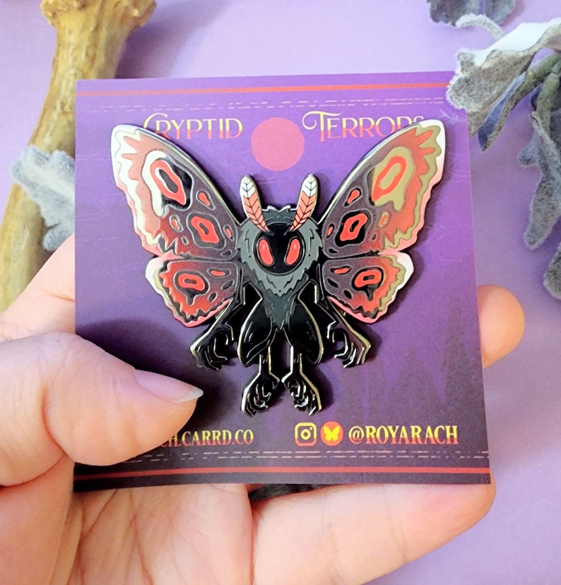 May include: A black and silver moth pin with red and tan wing details. The pin is on a purple card with the words "Cryptid Terrors" and social media handles. The moth has large eyes and a detailed body.