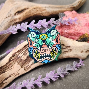 May include: A colorful enamel pin featuring a stylized, cartoonish monster face with a third eye, fangs, and swirling patterns. The pin is a vibrant mix of blue, green, pink, and yellow.