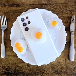 May include: A white smartphone case featuring a three-dimensional fried egg decoration, placed on a white scalloped plate with two additional fried egg ornaments, accompanied by a silver fork and a silver knife on either side.