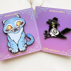 May include: Two enamel pins on purple backing cards. One pin is a light blue cat with dark blue stripes, yellow eyes, and sharp teeth. The other pin is a black and white bird wearing a hat with three yellow eyes. Text reads ROYARACH.CARRD.CO.
