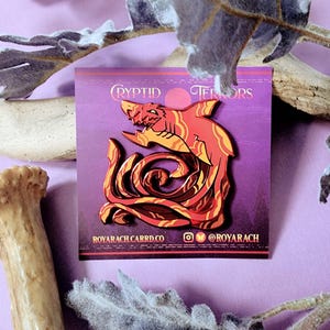 May include: A red and orange enamel pin depicting a shark with a swirling, leafy design. The pin reads "Cryptid Terrors" and "Royarach.carrd.co" with an Instagram icon and the handle "@royarach".
