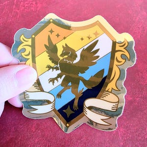 Puede incluir: Una pegatina dorada con forma de escudo, con un diseño de lobo alado. El escudo está dividido en secciones de naranja, amarillo, blanco, azul y marino. Hay pancartas decorativas en la parte inferior. La pegatina está sujeta por una mano.