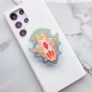 May include: A white smartphone with a colorful sticker featuring a winged creature design. The sticker has a light blue outline, orange wings, and a red heart-shaped center. The phone is on a white and gray marble surface.