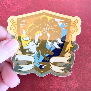 Puede incluir: Una pegatina con marco dorado con un diseño de escudo. El escudo presenta un zorro estilizado con una gran cola en forma de abanico, sobre un fondo azul, amarillo y naranja. Dos cintas blancas en la parte inferior. La pegatina tiene una superficie brillante y reflectante.