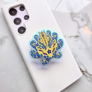 May include: A decorative phone grip featuring a colorful, abstract design. The grip has a light blue border with a yellow and blue central pattern. The phone grip is attached to a white phone case.