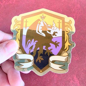 May include: A sticker featuring a shield design with a gold border and accents. The shield has a gold, purple, and black color scheme with a dragon and banner. The sticker is held by a hand.