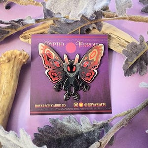 May include: A metal pin featuring a moth-like creature with red, black, and tan details. The pin is on a purple card with the text "Cryptid Terrors" and social media handles. The moth has large wings and a detailed body.