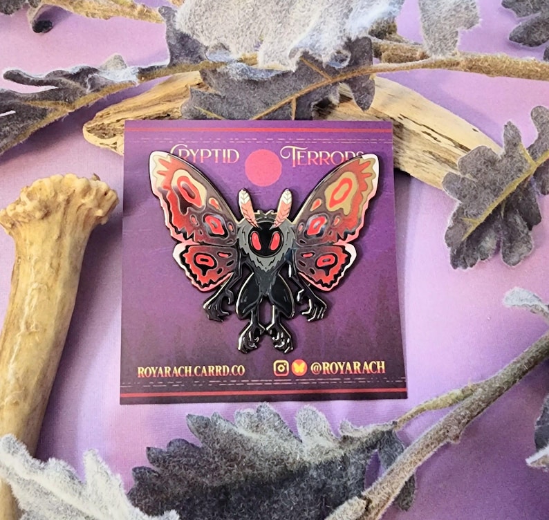 May include: A metal pin featuring a moth-like creature with red, black, and tan details. The pin is on a purple card with the text "Cryptid Terrors" and social media handles. The moth has large wings and a detailed body.