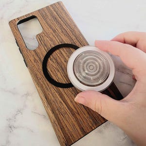 Może przedstawiać: Drewniane etui na telefon z czarnym okrągłym pierścieniem magnetycznym i przezroczystym uchwytem do telefonu z białą obwódką. Etui ma wzór słojów drewna. Uchwyt do telefonu jest trzymany w pobliżu etui.