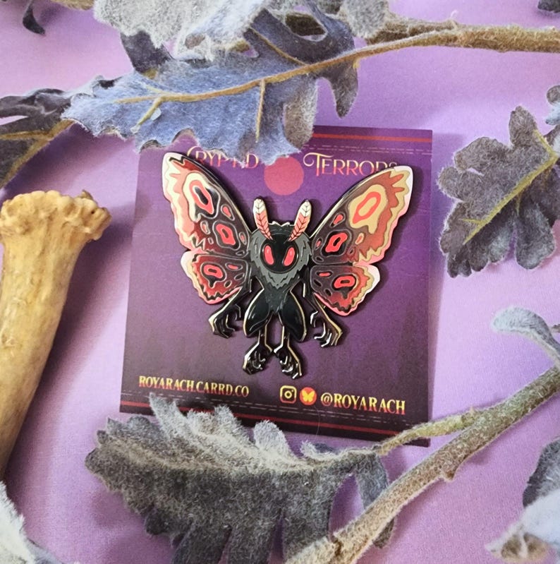 May include: A black and red moth pin with a detailed design, featuring a stylized moth with outstretched wings. The pin is set against a purple background with the text "DYPND TERRORS" and "ROYARACH.CARRD.CO".